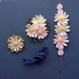 Könnte beinhalten: Drei gehäkelte Gänseblümchen-Blumenmuster in Weiß, Rosa und Grün. Die Muster sind auf einer blauen Oberfläche angeordnet. Das größte Muster ist ein Armband mit einer silbernen Kette. Die kleineren Muster sind Broschen.