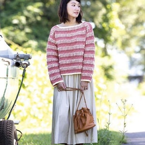 May include: A woman wearing a pink and white striped sweater with a white crocheted trim. She is also wearing a long, white skirt and a brown bucket bag.