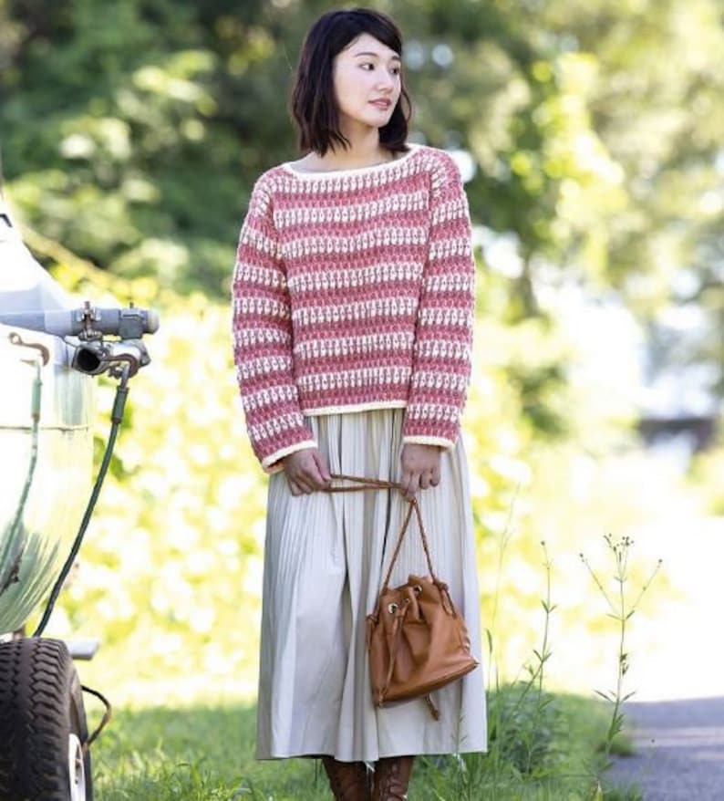 May include: A woman wearing a pink and white striped sweater with a white crocheted trim. She is also wearing a long, white skirt and a brown bucket bag.