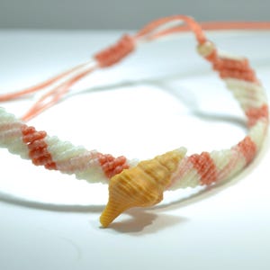 May include: A handmade macrame bracelet with alternating stripes of white, coral, and peach-colored threads. A small, light-brown seashell charm is attached to the bracelet. The bracelet is adjustable.