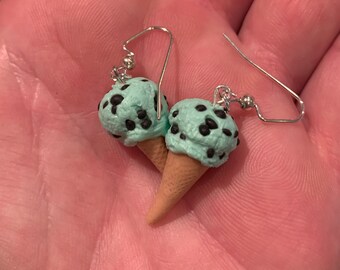 May include: A pair of novelty earrings featuring miniature mint chocolate chip ice cream cones. Each scoop of ice cream is a light green color with dark brown chocolate chips. The cones are a light brown color, and the earrings have silver-colored hooks.