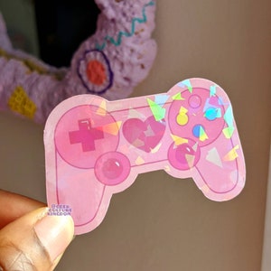 Puede incluir: Una pegatina rosa con forma de mando de videojuegos. El mando tiene una cruceta direccional, un corazón y varios botones. La pegatina tiene un acabado holográfico iridiscente. El texto "@GEEK CULTURE KINGDOM" es visible.