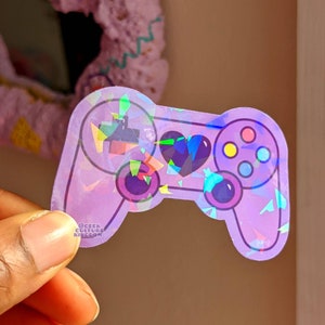 May include: A holographic purple video game controller sticker with a heart and a colorful button layout. The sticker has a glittery, iridescent finish and says "Geek Culture Kingdom" at the bottom.