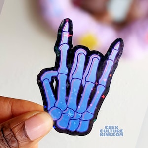 May include: A blue holographic sticker of a skeleton hand making the rock and roll hand gesture. The sticker has a black background with white stars. The sticker has the text "Geek Culture Kingdom" at the bottom.