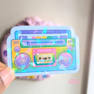 Può includere: Un adesivo olografico di un boombox retrò con un nastro cassette. Il boombox è giallo, rosa e blu con altoparlanti viola. L'adesivo ha uno sfondo blu chiaro con accenti olografici iridescenti.