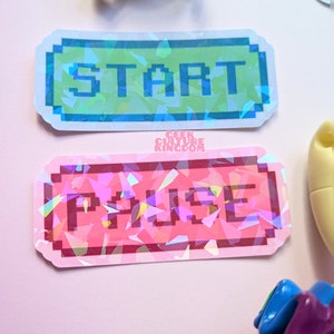 Könnte beinhalten: Zwei schimmernde holografische Aufkleber mit pixeligem Text. Der obere Aufkleber ist blau und grün mit dem Wort "START" in grün. Der untere Aufkleber ist rosa mit dem Wort "PAUSE" in rot.