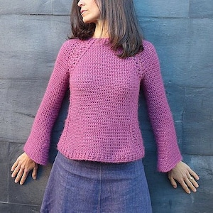 May include: A crocheted sweater in a deep purple shade with long sleeves and a round neckline. The sweater has a slightly flared silhouette and a delicate stitch pattern.