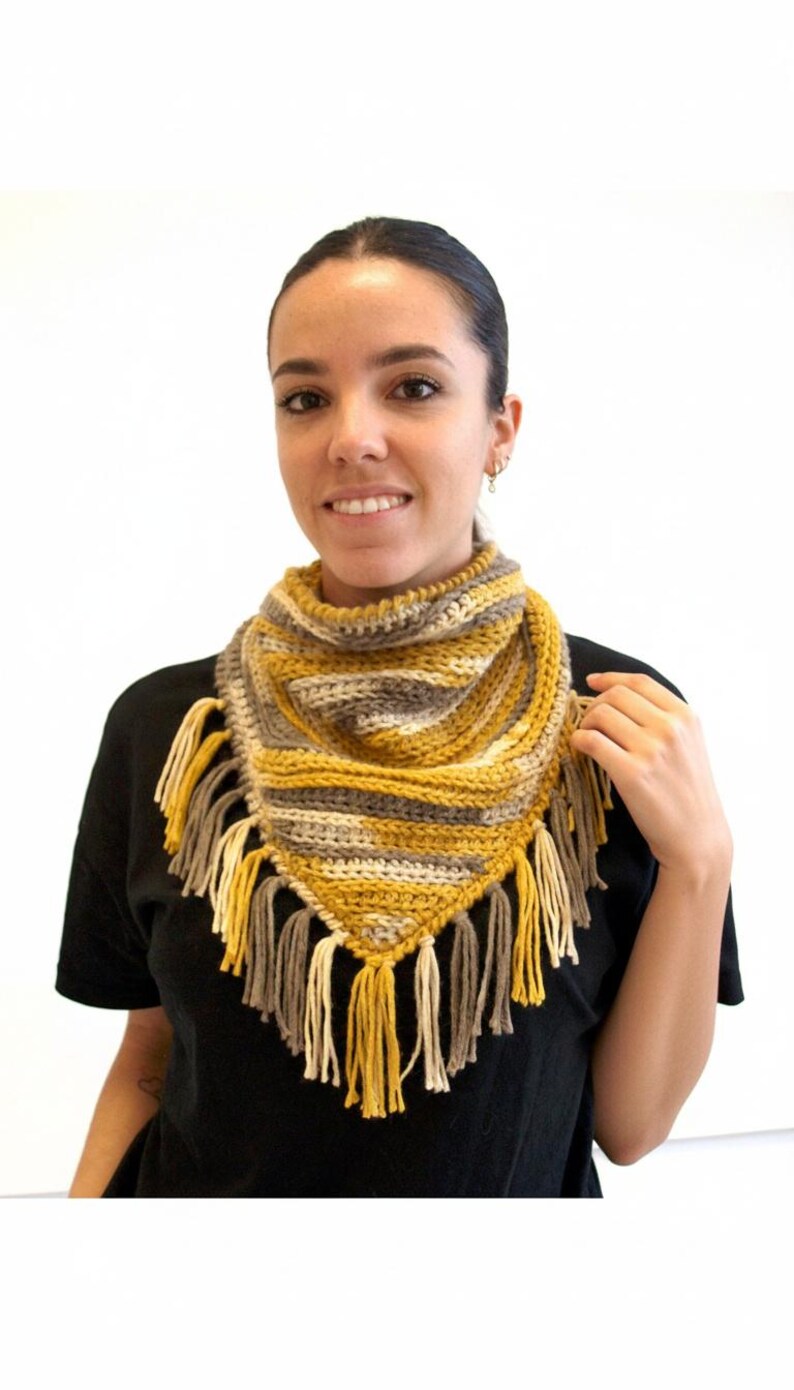 Puede incluir: Un pa&ntilde;uelo bandana triangular de ganchillo con rayas horizontales en amarillo mostaza, gris y crema. El accesorio est&aacute; rematado con largos y gruesos flecos de lana en los mismos tonos amarillo mostaza, gris y crema en el borde inferior.