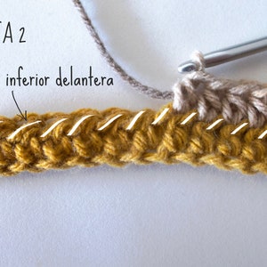 Puede incluir: Un proyecto de ganchillo en proceso con un ganchillo de mango azul, usando hilo amarillo mostaza y beige. El texto VUELTA 2 y Lazada inferior delantera es visible, con l&iacute;neas blancas que resaltan la lazada inferior delantera de los puntos.
