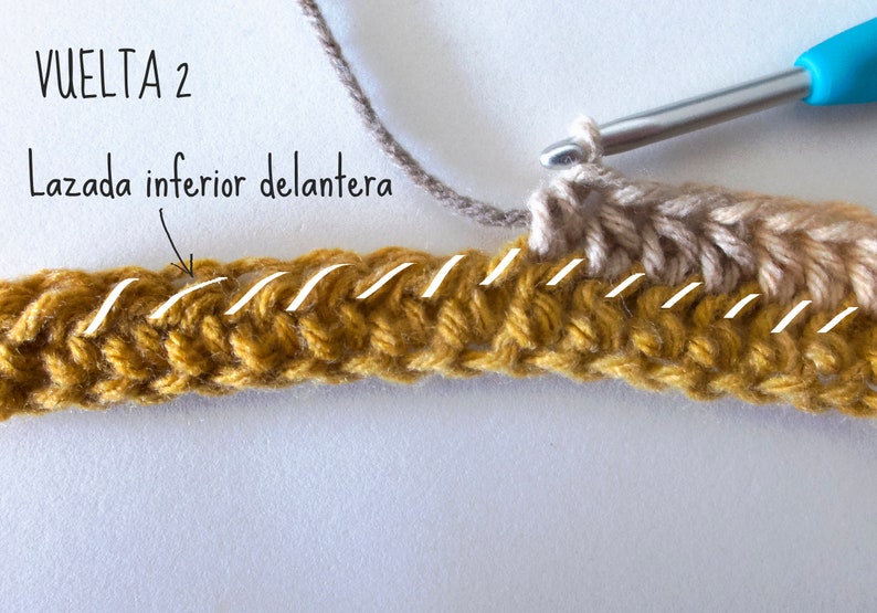 Puede incluir: Un proyecto de ganchillo en proceso con un ganchillo de mango azul, usando hilo amarillo mostaza y beige. El texto VUELTA 2 y Lazada inferior delantera es visible, con l&iacute;neas blancas que resaltan la lazada inferior delantera de los puntos.