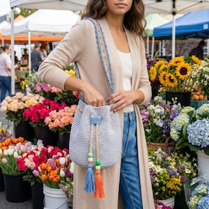 May include: A light blue crochet bucket bag with a drawstring closure and a colourful strap. The bag features blue and orange tassels with wooden beads. The strap has a rainbow trim. The bag is being held in front of a flower market.