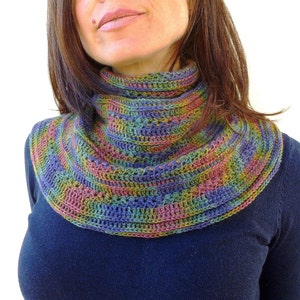 May include: A multicolored crochet cowl with a high neck. The cowl features a blend of blue, green, yellow, and pink yarns, creating a striped pattern. The garment is worn over a dark blue top.