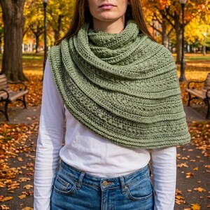 May include: A sage green crocheted shawl featuring a textured pattern with alternating rows of stitches, draped over a white long-sleeve shirt and blue denim jeans. The shawl has a wide, layered design that wraps around the shoulders and neck.