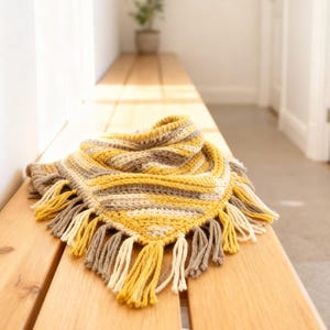 Puede incluir: Una bufanda triangular de ganchillo con rayas horizontales en amarillo mostaza, crema y gris claro. La bufanda est&aacute; rematada con un fleco decorativo a lo largo del borde inferior, compuesto por borlas de hilo alternas en la misma paleta.