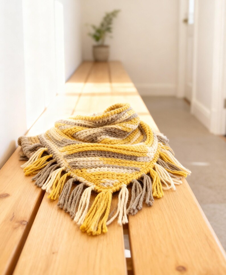 Puede incluir: Una bufanda triangular de ganchillo con rayas horizontales en amarillo mostaza, crema y gris claro. La bufanda est&aacute; rematada con un fleco decorativo a lo largo del borde inferior, compuesto por borlas de hilo alternas en la misma paleta.