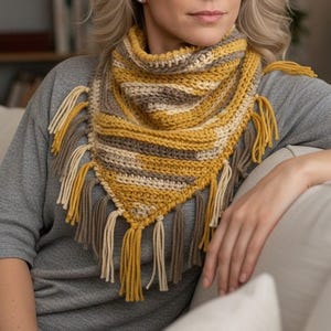 Puede incluir: Un pa&ntilde;uelo triangular de ganchillo con rayas horizontales en amarillo mostaza, crema y topo. La bufanda tiene una textura acanalada y est&aacute; rematada con largos y gruesos flecos de hilo a lo largo de los bordes inferiores en tonos a juego.
