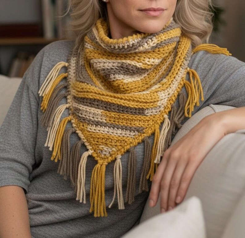 Puede incluir: Un pa&ntilde;uelo triangular de ganchillo con rayas horizontales en amarillo mostaza, crema y topo. La bufanda tiene una textura acanalada y est&aacute; rematada con largos y gruesos flecos de hilo a lo largo de los bordes inferiores en tonos a juego.