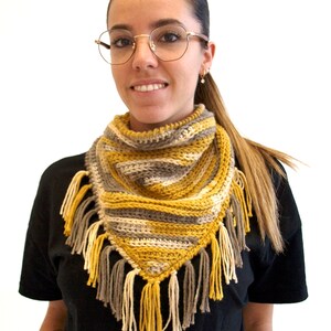 Puede incluir: Un pa&ntilde;uelo triangular de ganchillo con rayas horizontales en amarillo mostaza, gris y crema. El accesorio est&aacute; rematado con un fleco decorativo en el borde inferior, compuesto por borlas de hilo alternas en colores a juego.