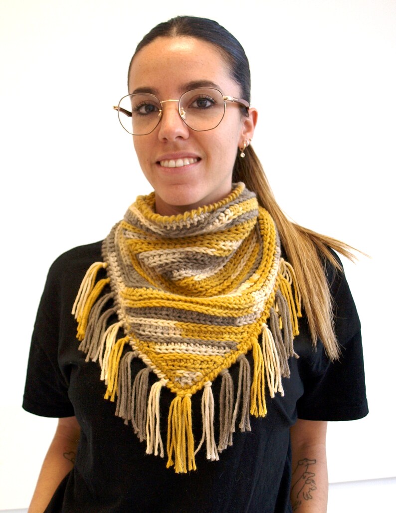 Puede incluir: Un pa&ntilde;uelo triangular de ganchillo con rayas horizontales en amarillo mostaza, gris y crema. El accesorio est&aacute; rematado con un fleco decorativo en el borde inferior, compuesto por borlas de hilo alternas en colores a juego.