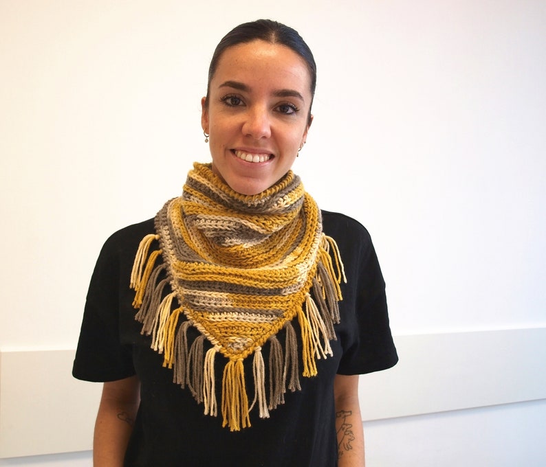 Puede incluir: Un pa&ntilde;uelo triangular de ganchillo con rayas horizontales en amarillo mostaza, crema y gris oscuro. El accesorio est&aacute; rematado con largos y gruesos flecos de hilo a lo largo de los bordes inferiores, creando un aspecto texturizado y superpuesto.