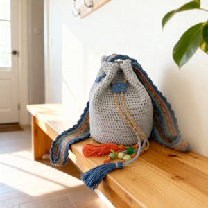Könnte beinhalten: Eine hellgraue Häkel-Beuteltasche mit Kordelzugverschluss und geflochtenen Riemen in Orange, Grün und Blau. Die Tasche verfügt über lange, dekorative Seitenteile mit gewellten Rändern aus mehrfarbigem Garn sowie hängende Quasten mit Holzperlen.