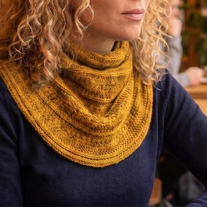 May include: A mustard yellow knitted cowl scarf featuring a textured pattern with horizontal ribbed bands and intricate stitch details. The scarf is draped around the neck and rests over a dark navy blue long-sleeved shirt.