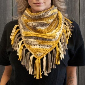 May include: A triangular crochet cowl scarf featuring a variegated pattern of mustard yellow, cream, and taupe yarn. The accessory has a ribbed texture and is finished with long, thick yarn fringe tassels along the bottom edges and point.
