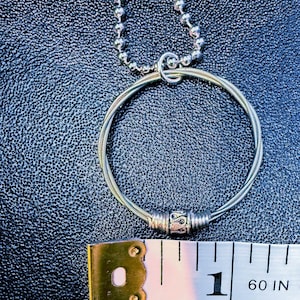 May include: A silver-toned necklace featuring a circular pendant made from coiled wire, with a decorative bead. The necklace is on a textured black surface, with a measuring tape showing 1 inch.