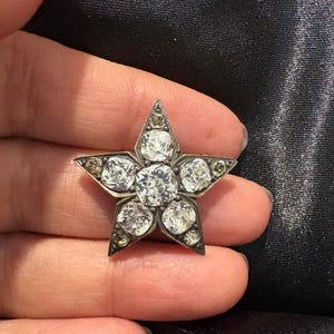 May include: A star-shaped brooch with a silver-tone metal frame. The brooch is adorned with clear, round-cut gemstones, with a larger stone at the center. The brooch is held in a hand against a dark background.