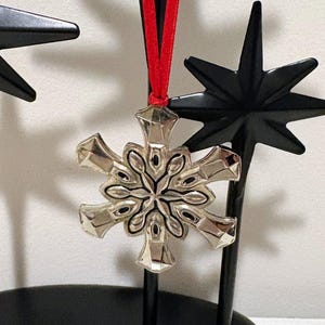 May include: A silver snowflake ornament with a red ribbon for hanging. The snowflake has a detailed design with black accents. It is displayed near black star-shaped decorations, creating a festive holiday theme.