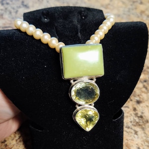 May include: A necklace featuring a strand of cream-colored pearls. The pendant has a large, light green rectangular stone at the top, with two yellow gemstones below. The pendant is set in silver. The necklace is displayed on a black velvet stand.