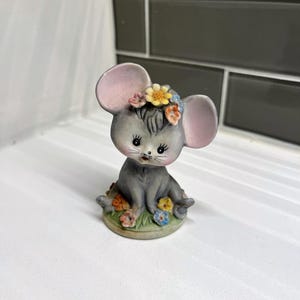 May include: A small gray ceramic figurine of a mouse wearing a flower crown. The mouse is sitting on a green base with flowers.