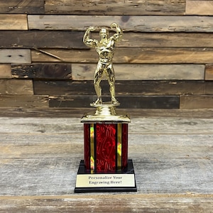 May include: A gold-colored trophy featuring a weightlifter figure atop a red and gold base. The base has a black bottom with a gold plate that reads "Personalize Your Engraving Here!" The trophy is set against a wooden background.