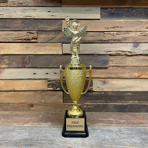 May include: A gold trophy with a winged figure holding a diploma on top. The base of the trophy is black with the words "FREE ENGRAVING" on a gold plate.