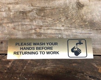 May include: A rectangular, gold-colored metal sign with black text that reads "PLEASE WASH YOUR HANDS BEFORE RETURNING TO WORK." A black and white graphic of hands under a faucet is on the right side.