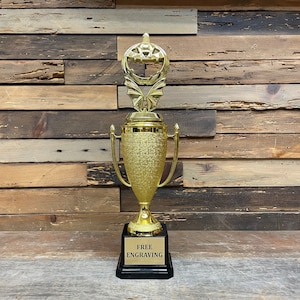 May include: A gold-colored trophy with a pumpkin-shaped top. The base of the trophy has a black plaque with the words "FREE ENGRAVING" on it.