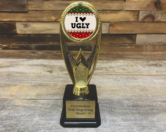 以下が含まれることがあります： 「I ♥ UGLY」という文字が書かれた、赤、緑、白のパターンが施された丸いトップを持つ金色のトロフィー。トロフィーのベースは黒で、金色のプレートには「刻印をカスタマイズしてください！」と書かれています。