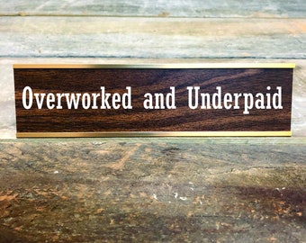May include: A brown desk nameplate with a gold trim and the text "Overworked and Underpaid" in white.