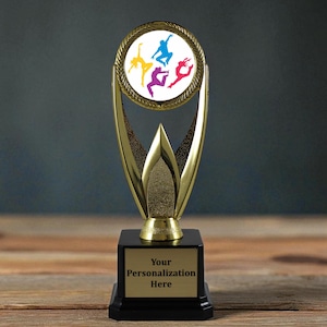 May include: A gold-colored trophy with a black base and a circular emblem featuring colorful dancing figures. The trophy has a space for personalization. The background is a blurred wooden surface.