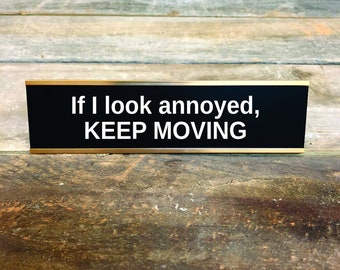May include: A black desk sign with gold trim features the white text: "If I look annoyed, KEEP MOVING." The sign is set against a weathered, wooden background.