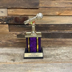 May include: A gold trophy featuring a microphone. The trophy has a purple and gold base, with the words "FREE ENGRAVING" on a black plate.
