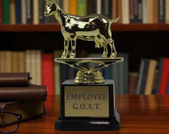 May include: A gold-colored goat trophy with the text "EMPLOYEE G.O.A.T." on a black base. The trophy is on a wooden surface with books and a pair of glasses in the background. The goat figurine is the focal point.