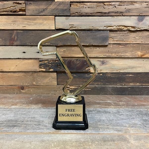 May include: Gold trophy shaped like a race track with a black base. The base has a gold plaque that says "FREE ENGRAVING".