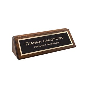 May include: A wooden desk nameplate with a black nameplate and gold trim. The nameplate reads "DIANNA LANGFORD, PROJECT MANAGER" in white text. The nameplate is made of dark brown wood and has a triangular shape.