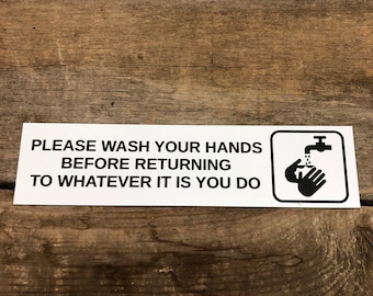 May include: White sign with black text that reads "Please wash your hands before returning to whatever it is you do". A black and white icon of a hand washing under a faucet is on the right side of the sign.