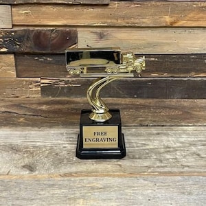 Puede incluir: Un trofeo dorado con un diseño de camión. El trofeo tiene una base negra con una placa dorada que dice "FREE ENGRAVING". El camión está detallado con ruedas y un remolque, sobre un fondo de madera.