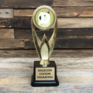 Może przedstawiać: Złote trofeum z czarną podstawą. Trofeum posiada logo Dogecoin w okrągłej ramce na górze. Podstawa ma złotą płytkę z napisem "DOGECOIN CUSTOM ENGRAVING".