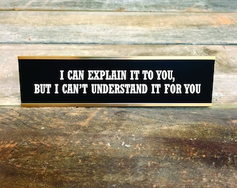 May include: A black desk nameplate with gold trim features the white text: "I CAN EXPLAIN IT TO YOU, BUT I CAN'T UNDERSTAND IT FOR YOU." The nameplate rests on a weathered wooden surface.