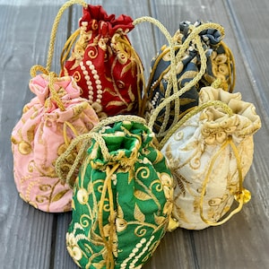 May include: A collection of five embroidered drawstring pouches in various colors: red, pink, green, gray, and cream. Each pouch features intricate gold floral embroidery and a gold cord drawstring closure. The pouches are arranged on a gray wooden surface.