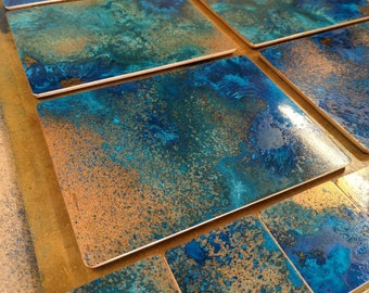 May include: A collection of rectangular and square decorative tiles with a copper-colored border. The tiles feature a vibrant, abstract design with shades of blue, teal, and bronze, creating a textured, artistic appearance. The tiles are arranged on a light-colored surface.